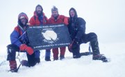 Group of men in winter equipment in a snowy landscape holding up a board with Antarctica on it.