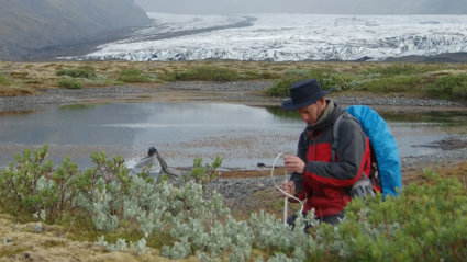 Person setting up a measuring device in front of a glacier and lake. The person is wearing a hat and rucksack.