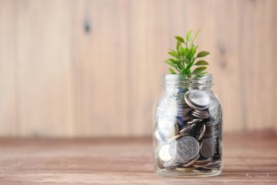 A glass jar filled with silver coins, from which a green plant emerges on top on top sits on a wooden surface 