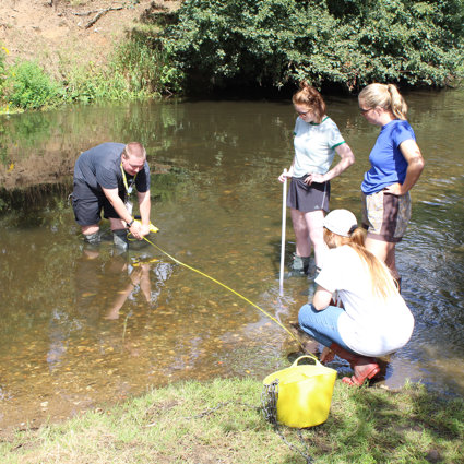 A grop of teachers are standing in a shallow river, two are holding a tape measure across the river to measure its width. Two are standing by and watching.