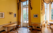 A room with yellow wallpapers and curtains. There are sets of tables and chairs and hung portraits, the garden is visible through the windows