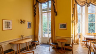 A room with yellow wallpapers and curtains. There are sets of tables and chairs and hung portraits, the garden is visible through the windows