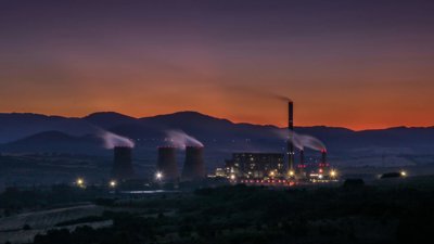 Industrial power plant at dusk, with cooling towers releasing steam against a mountain skyline.