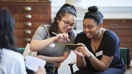 Two young people sat smiling while looking at a phone together. In the background are large woodend map drawers.