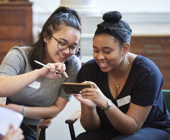 Two young people sat smiling while looking at a phone together. In the background are large woodend map drawers.