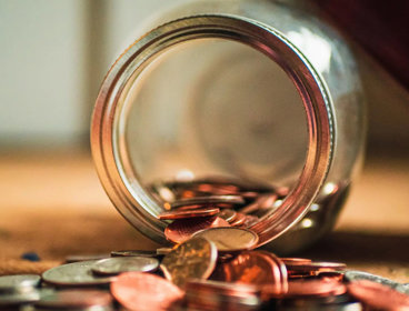 Close-up photo of assorted coins coming out of a glass jar laying on its side.