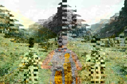 A person wearing a backpack seen from behind standing on an unpaved path surounded by grass overlooking mountains.