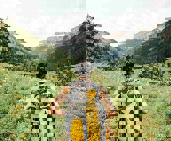 A person wearing a backpack seen from behind standing on an unpaved path surounded by grass overlooking mountains.