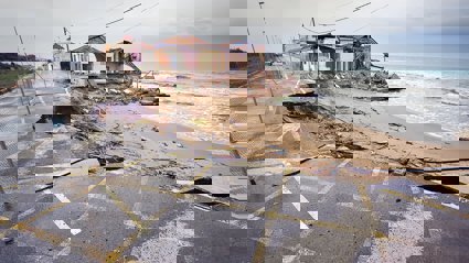 A damaged house and road by the coast. Rubble lays on the beach.