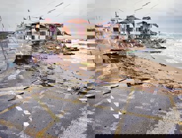 A damaged house and road by the coast. Rubble lays on the beach.