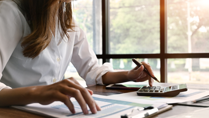 A person in a white shirt sits a a wooden desk whilst typing on a calculator and looking at paper on a clip board. the paper features charts and tables.
