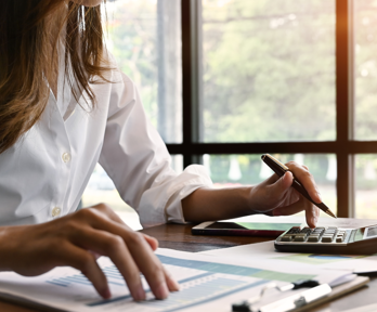 A person in a white shirt sits a a wooden desk whilst typing on a calculator and looking at paper on a clip board. the paper features charts and tables.