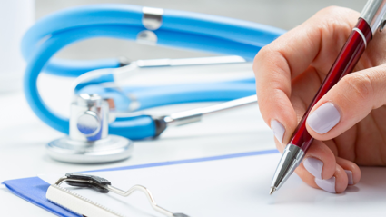 A person uses a pen to write on a peice of paper attached to a clip board. a stethoscope sits on the table next to the paper and clip board
