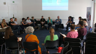 Group of people sit in a circle at a conference, they are sitting on black chairs.