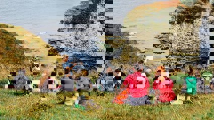 A group of young people sit on a seaside cliff, listening to a presenter in the front who's writing on a paper pad.