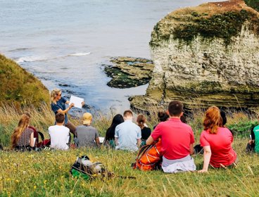 A group of young people sit on a seaside cliff, listening to a presenter in the front who's writing on a paper pad.