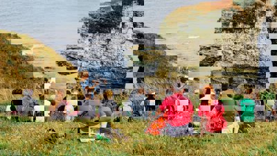 A group of young people sit on a seaside cliff, listening to a presenter in the front who's writing on a paper pad.