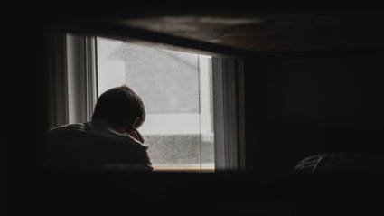 Person faced away from the viewer looks outside of the window whilst their head rests on their hand. The room around them is dark and the landscape ouside the window is grey and blurred.