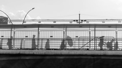 Silhouettes of people walking along a fenced pedestrian bridge against a bright sky in black and white.