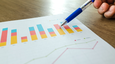 A bar chart and line graph printed are printed on a piece of paper which sits on a wooden desk. A person holds a pen and uses it to point to the bar chart near the top of the page.