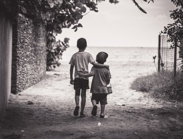 Two children with their arms around one another walking towards a beach