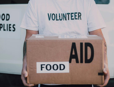 Person in a volunteer shirt holding a food aid box in front of a van marked food supplies.