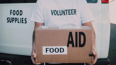 Person in a volunteer shirt holding a food aid box in front of a van marked food supplies.