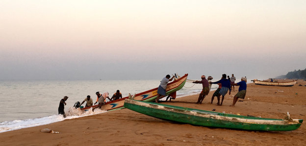 Two fishing boats on a beach pictured with people on either side pulling it out of the water. 