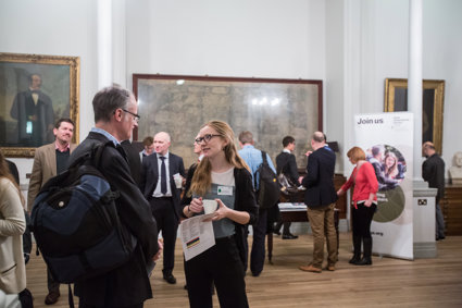 People stand scattered throughput the Royal Society map room. Most people appear to be mid conversation. Maps and portraits fill the walls behind.