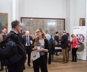 People stand scattered throughput the Royal Society map room. Most people appear to be mid conversation. Maps and portraits fill the walls behind.