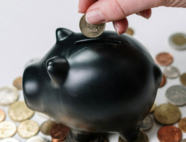 Black piggy bank surrounded by coins, with a hand adding a coin into the slot.