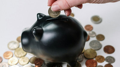 Black piggy bank surrounded by coins, with a hand adding a coin into the slot.