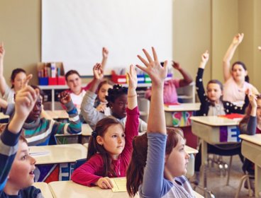 A classroom full of children, all raising a hand or finger in the air.