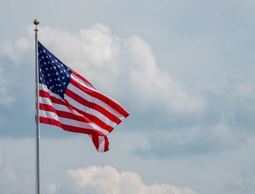 Flag of the USA on a pole waving against cloudy backdrop.