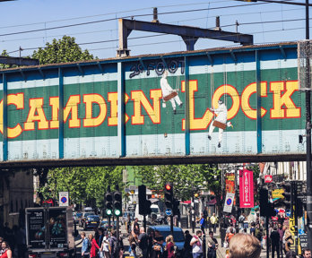 A railway bridge in Camden in London, with Camden Lock painted onto the side.  There are people below the bridge crossing a busy road and heading towards local shops