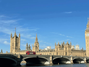 Houses of Parliament in London under a blue sky, with a red double-decker bus crossing Westminster Bridge in front.