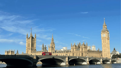 Houses of Parliament in London under a blue sky, with a red double-decker bus crossing Westminster Bridge in front.