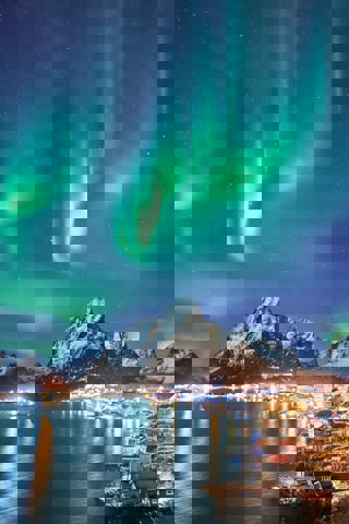 A view of a coastal town with a mountain backdrop, with the northern lights shiig green overhead