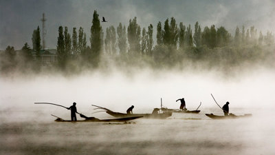 A misty lage with four boats and fishermen, a bird flying overhead in the distance.