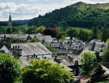 Rooftops of houses in a village with a church spire and hills with trees in the background. 