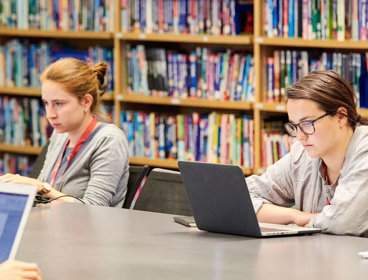 People sitting at a desk, working on laptops in a reading room library.