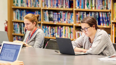 People sitting at a desk, working on laptops in a reading room library.