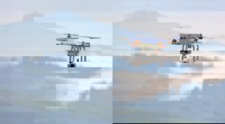 A grey and orange drone flying in the sky, a blurry background of forested hills can be seen