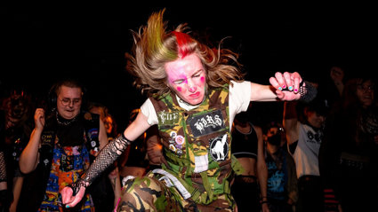 Person with face paint and dyed hair mid-dance in a dark venue, wearing a patch-covered camouflage vest, with a crowd behind.
