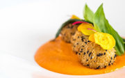Three sesame-coated fried balls on orange sauce, topped with yellow crisps and greens on a white plate.