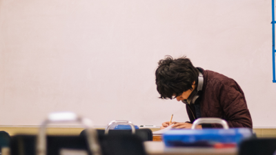 A high school student sits a fair distance away at a desk whilst writing on a sheet of paper. A plain white board fills the wall behind them.