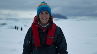 Lucy Obolensky in winter wear in the Arctic. The background is blurred and shows a plain of ice and contours of three penguins.