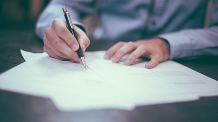 A person writes on a sheet of paper sticking out from a shallow stack of papers on top of a dark brown surface. The papers and background are blurred whist only the hands of the person can be seen