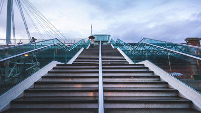 Modern city stairs shot from below. One person is at the top of the stairs, walking down and holding an umbrella. 