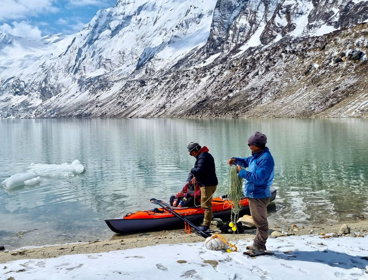 Two researchers on a lakeshore loading submersible temperature sensor arrays into a kayak, with snowclad Nepalese mountains in the background.